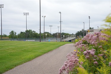 A paved trail line by flowers leading up to a ballfield