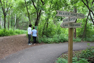 Two people read a trail map, behind them is a sign with arrows pointing in different directions with locations written on it 
